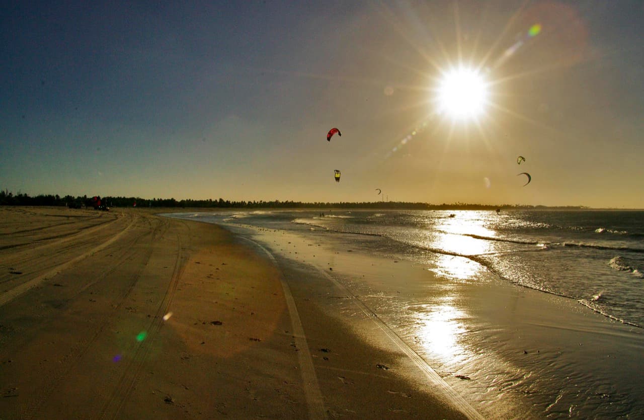 Kites no ar sobre a faixa de areia de São Miguel do Gostoso no fim de tarde, com o sol baixo refletindo na água rasa